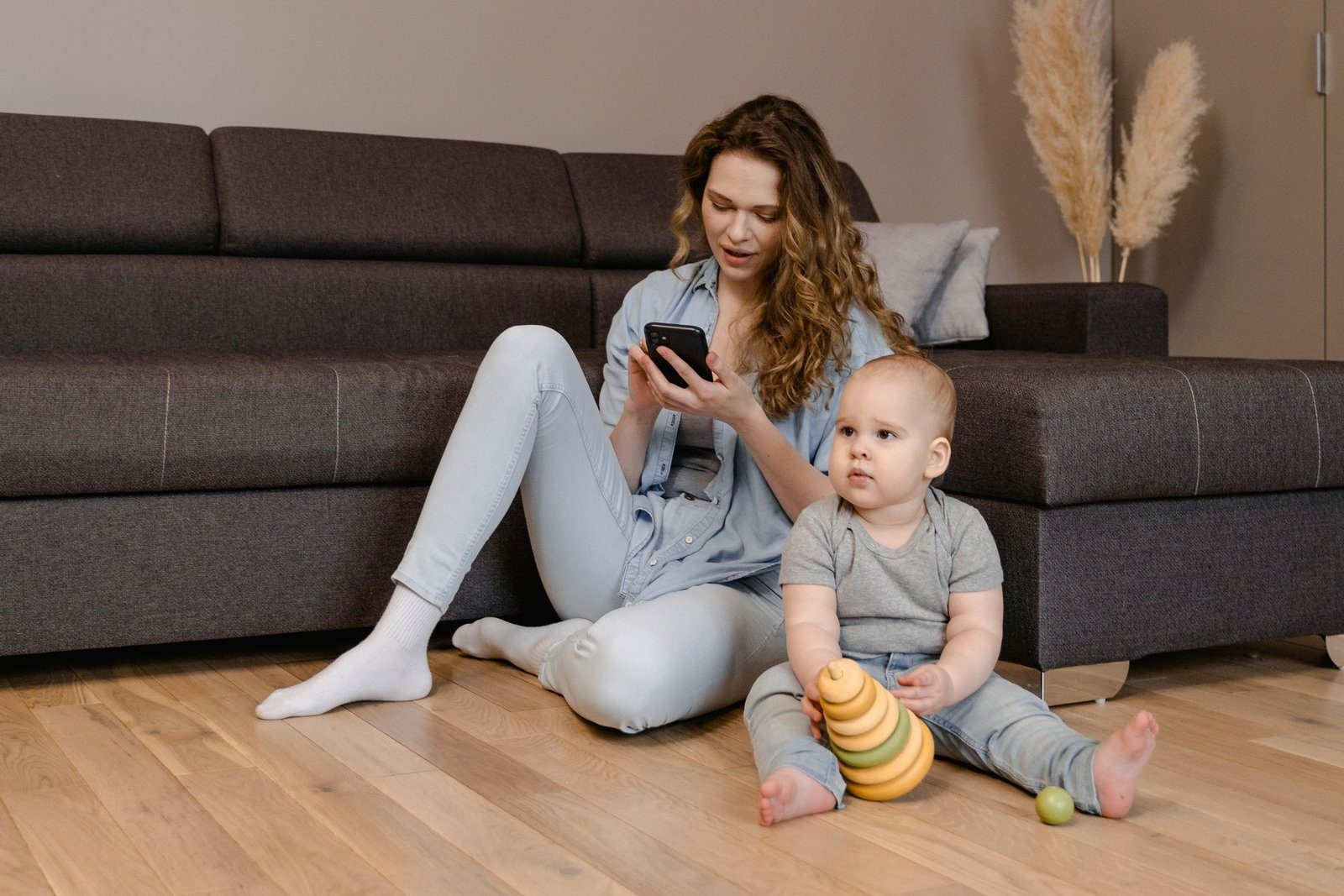 A mother sitting on the living room floor checking her phone while her baby plays with stacking rings