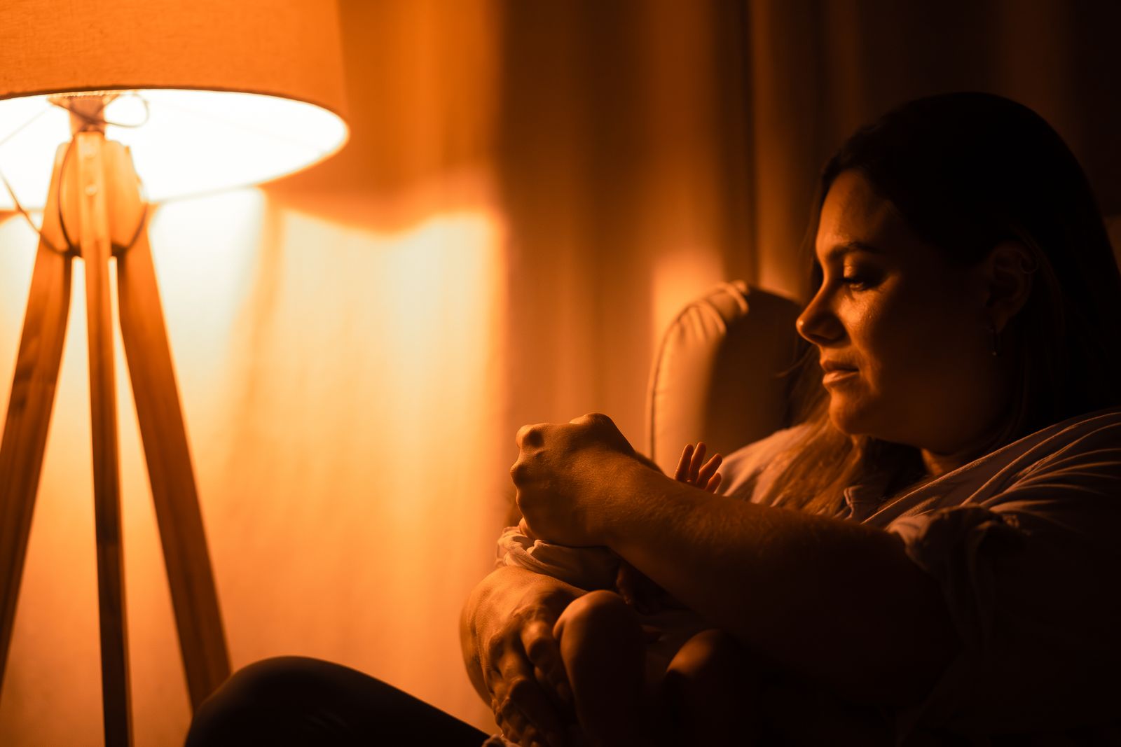 A mother holding her baby in a chair beside a glowing lamp during an evening feed