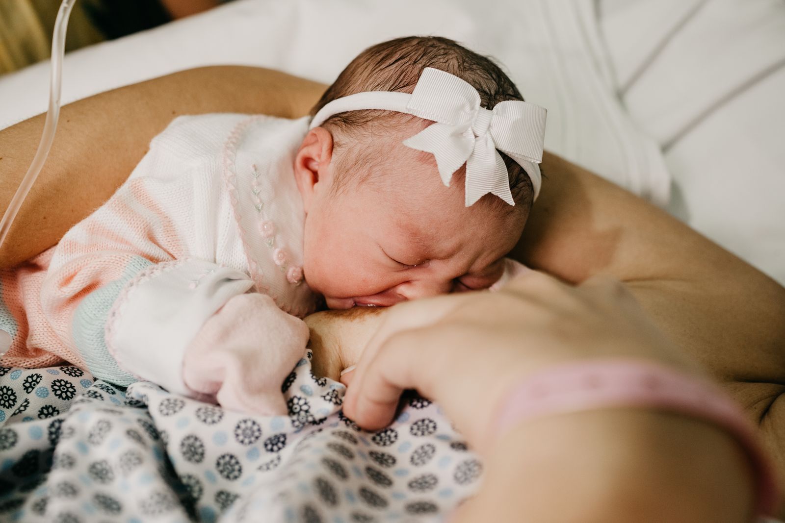 A newborn in a white hair bow nursing while resting on her mother's arm