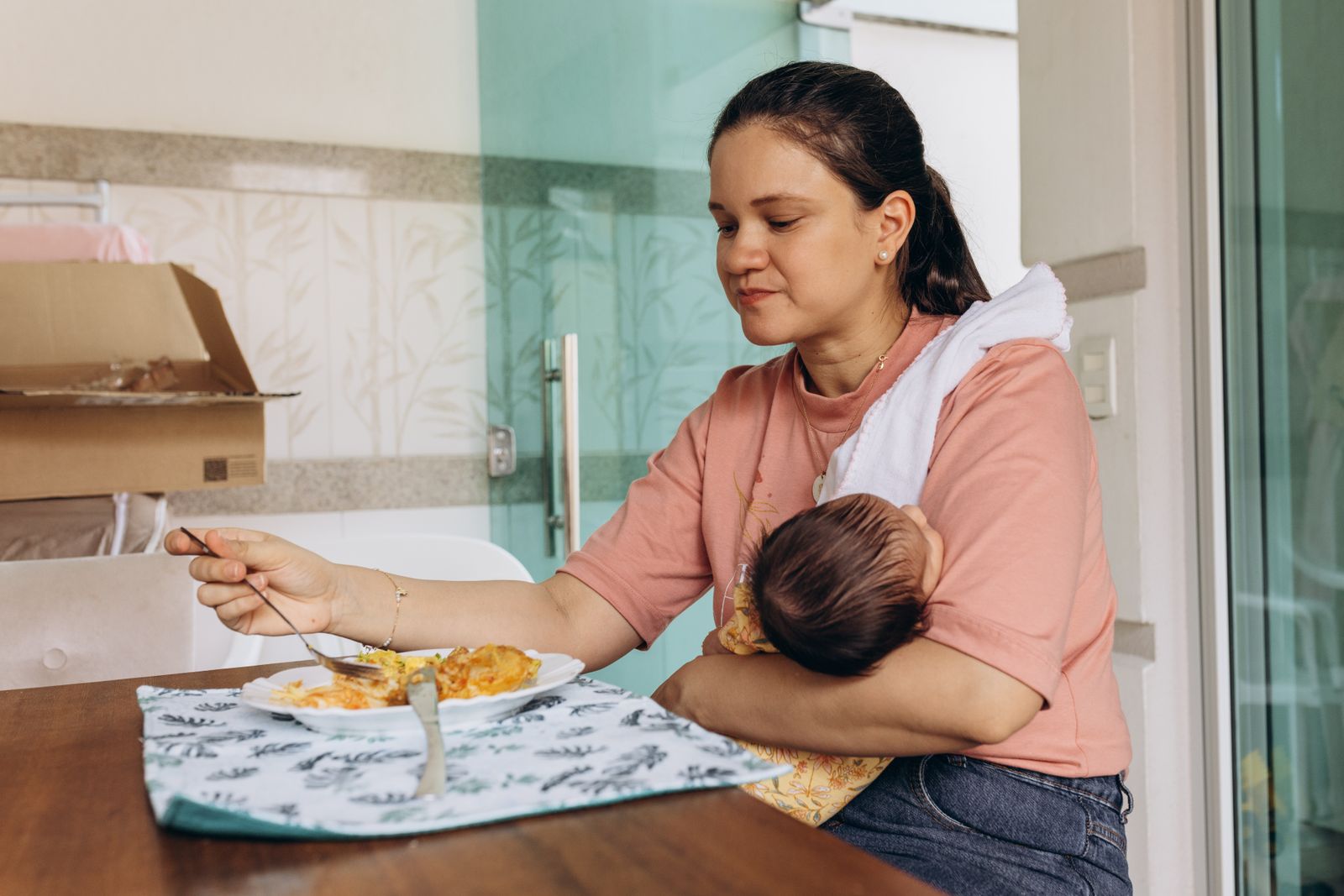 A mother eating a hot meal at the table with her baby held in one arm