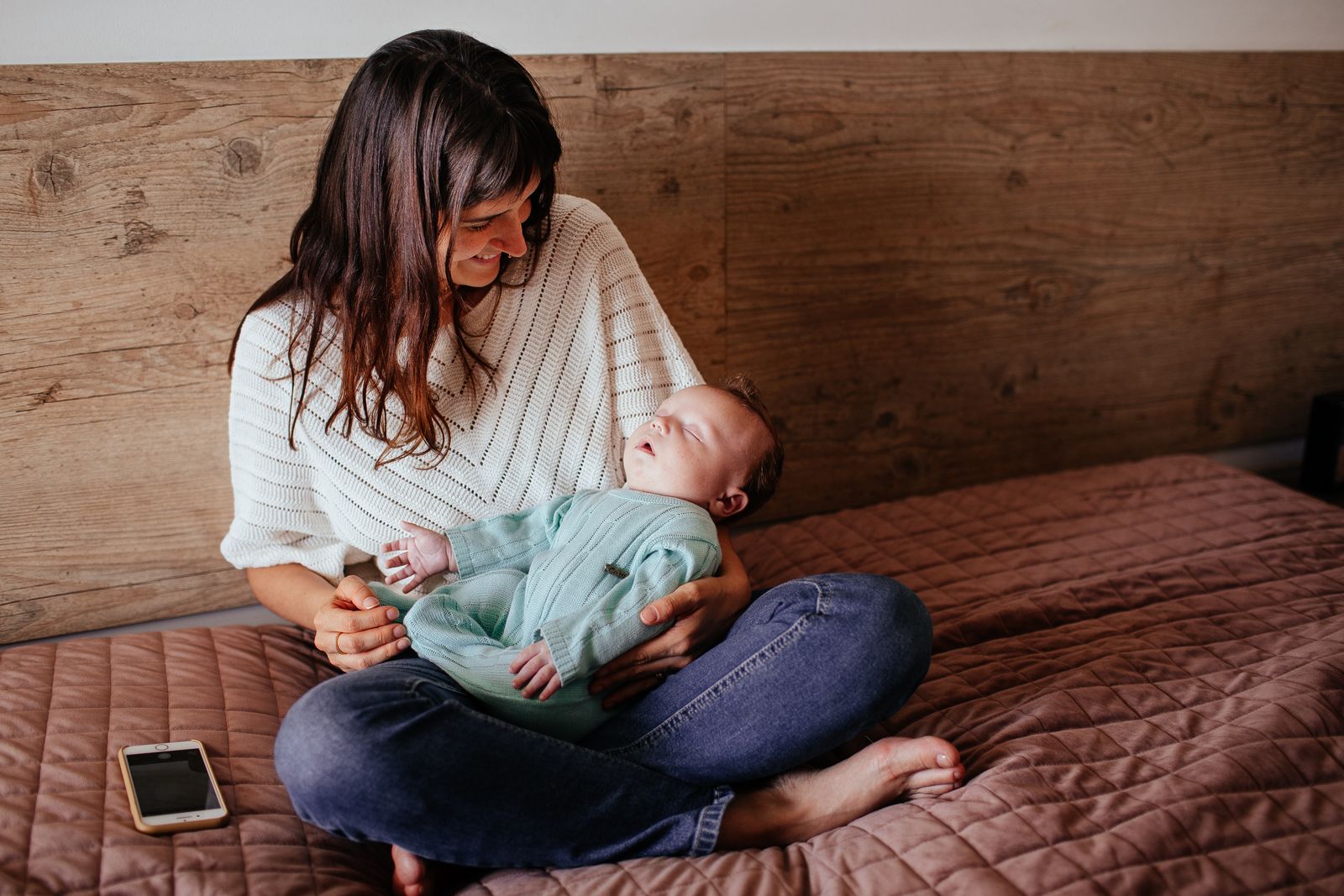A mother smiling down at her baby on a bed with a phone resting beside her