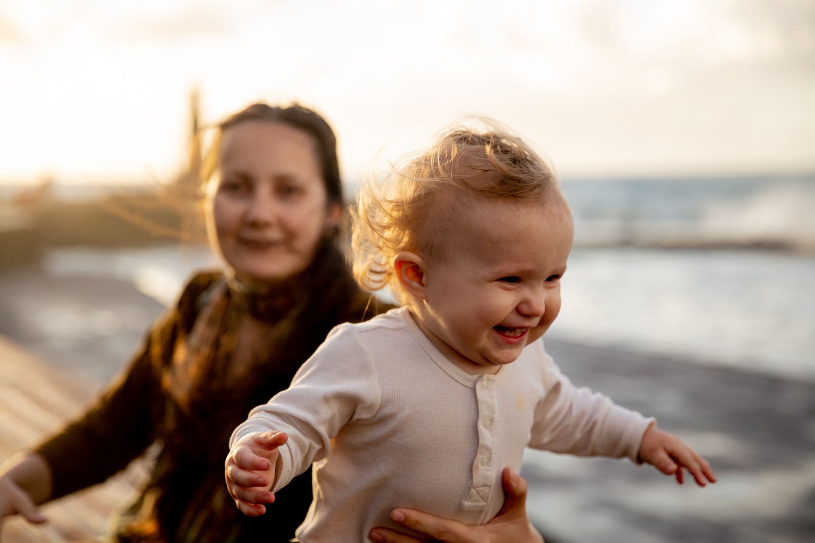 A laughing, rosy-cheeked baby with their mother smiling in the background by the water