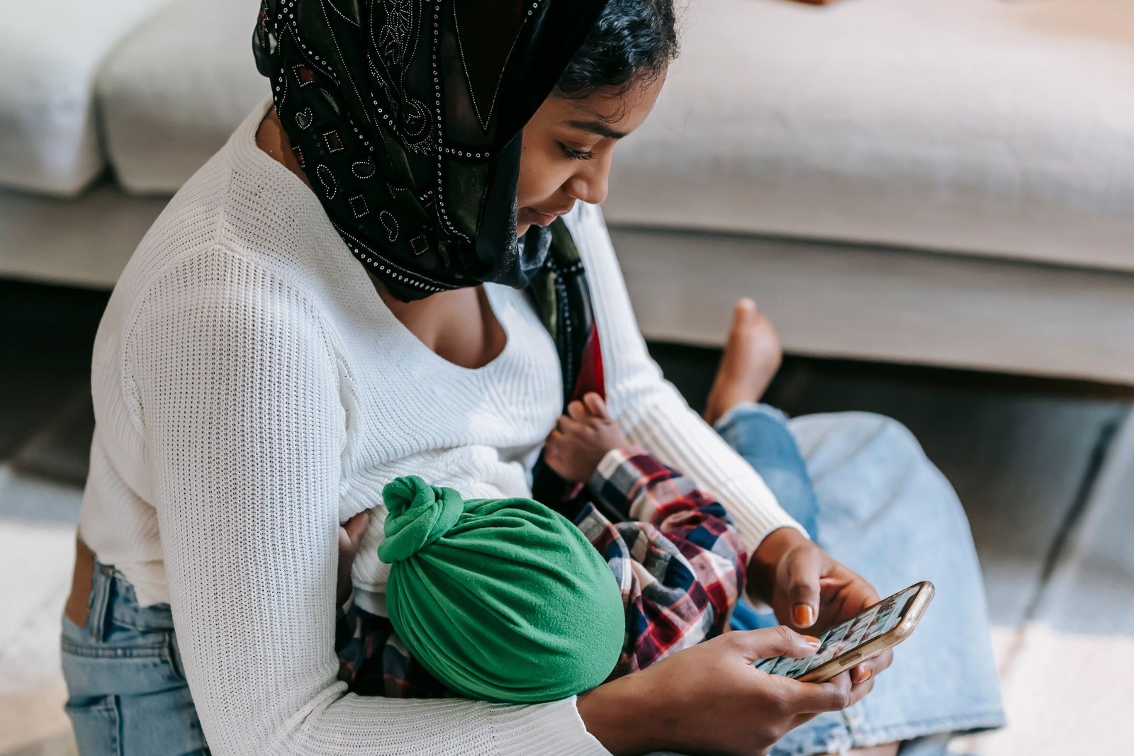 A mother sitting on the floor nursing her baby while looking at her phone