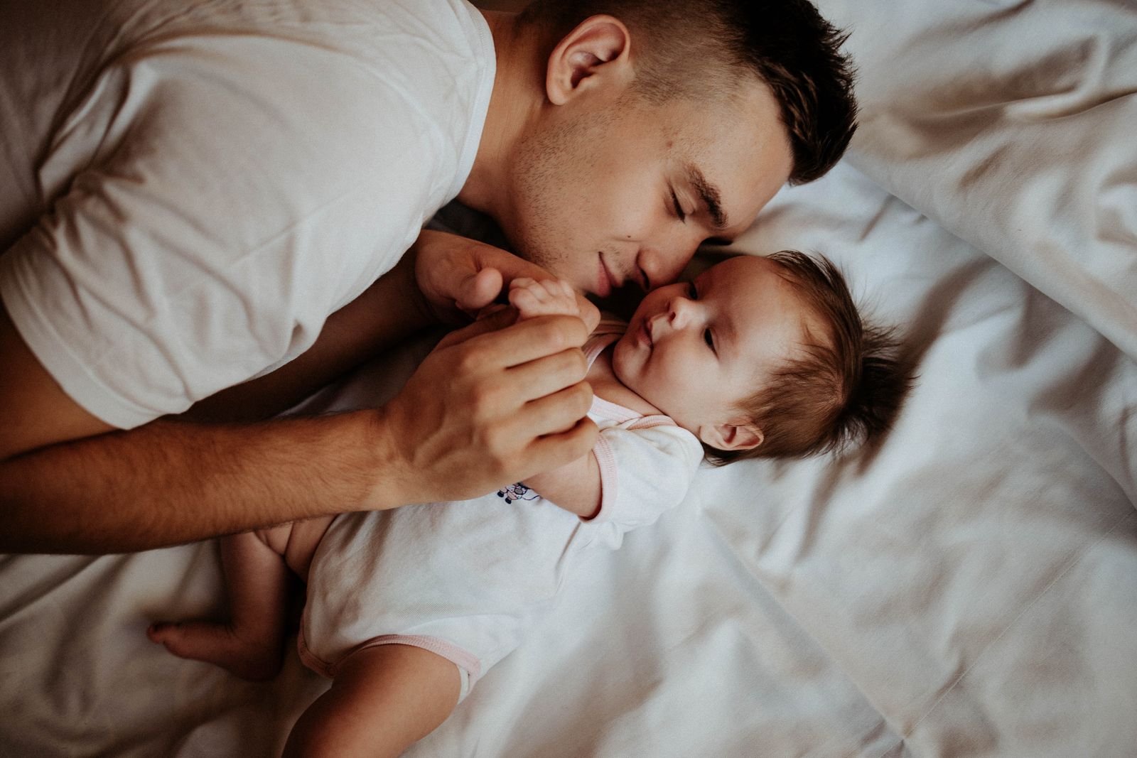 A father lying on white sheets nose to nose with his newborn baby