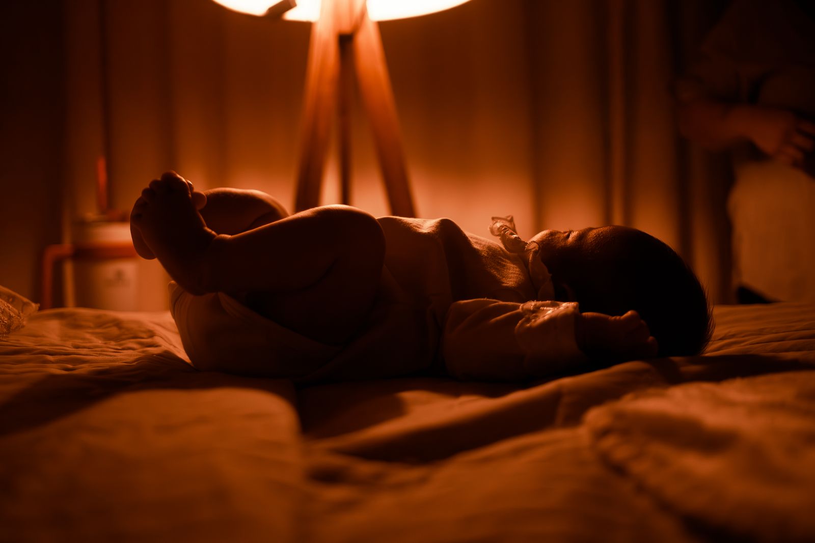 A baby asleep on a bed at night, lit by the warm glow of a bedside lamp